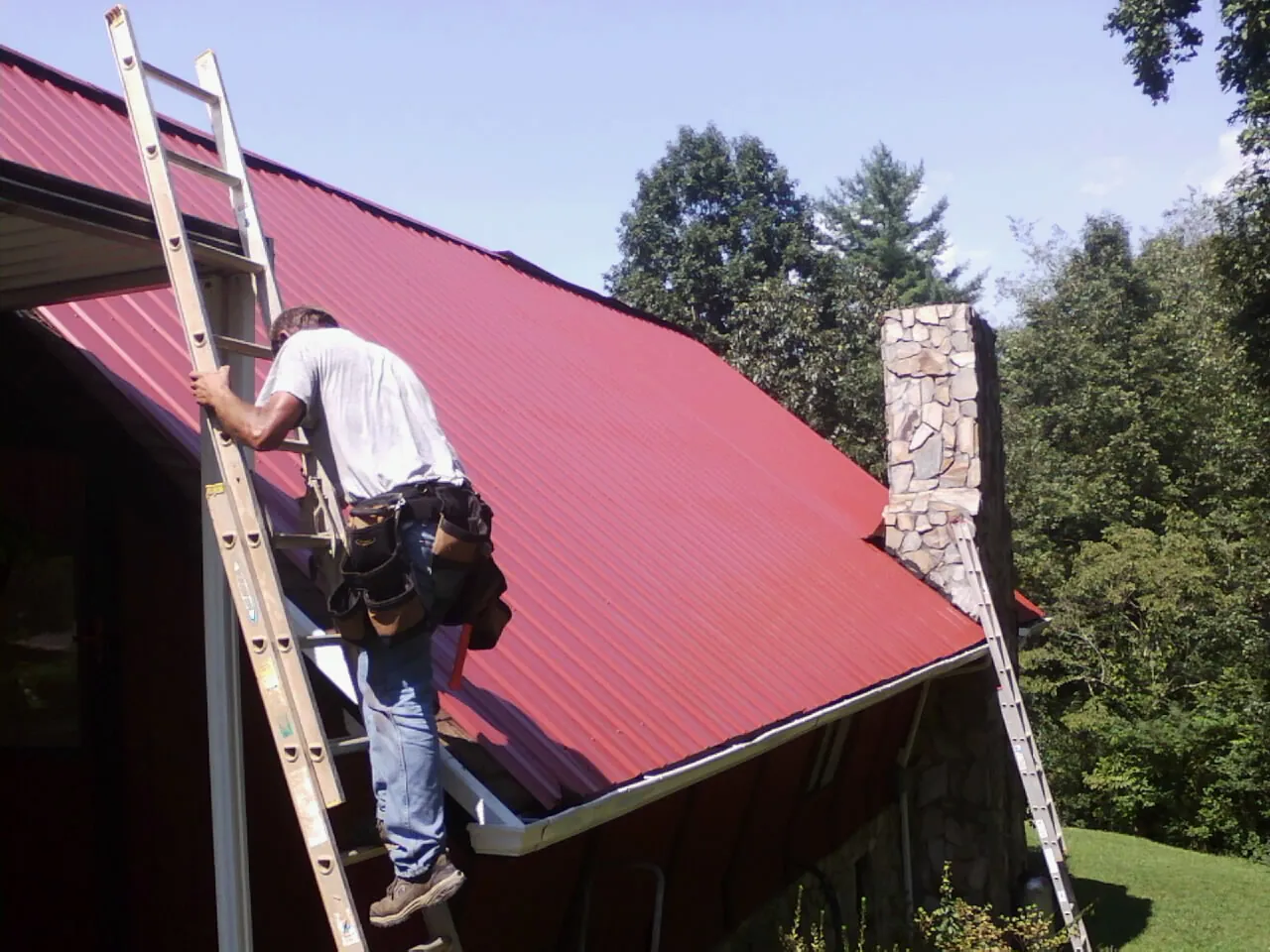 Roofer on a ladder beside a red metal roof and stone chimney — Soffit & Fascia Repair by Roofing Brothers in Filley